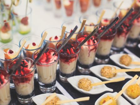 Assortment of desserts in glasses topped with fruit and skewers, alongside small plates of round appetizers with sticks.