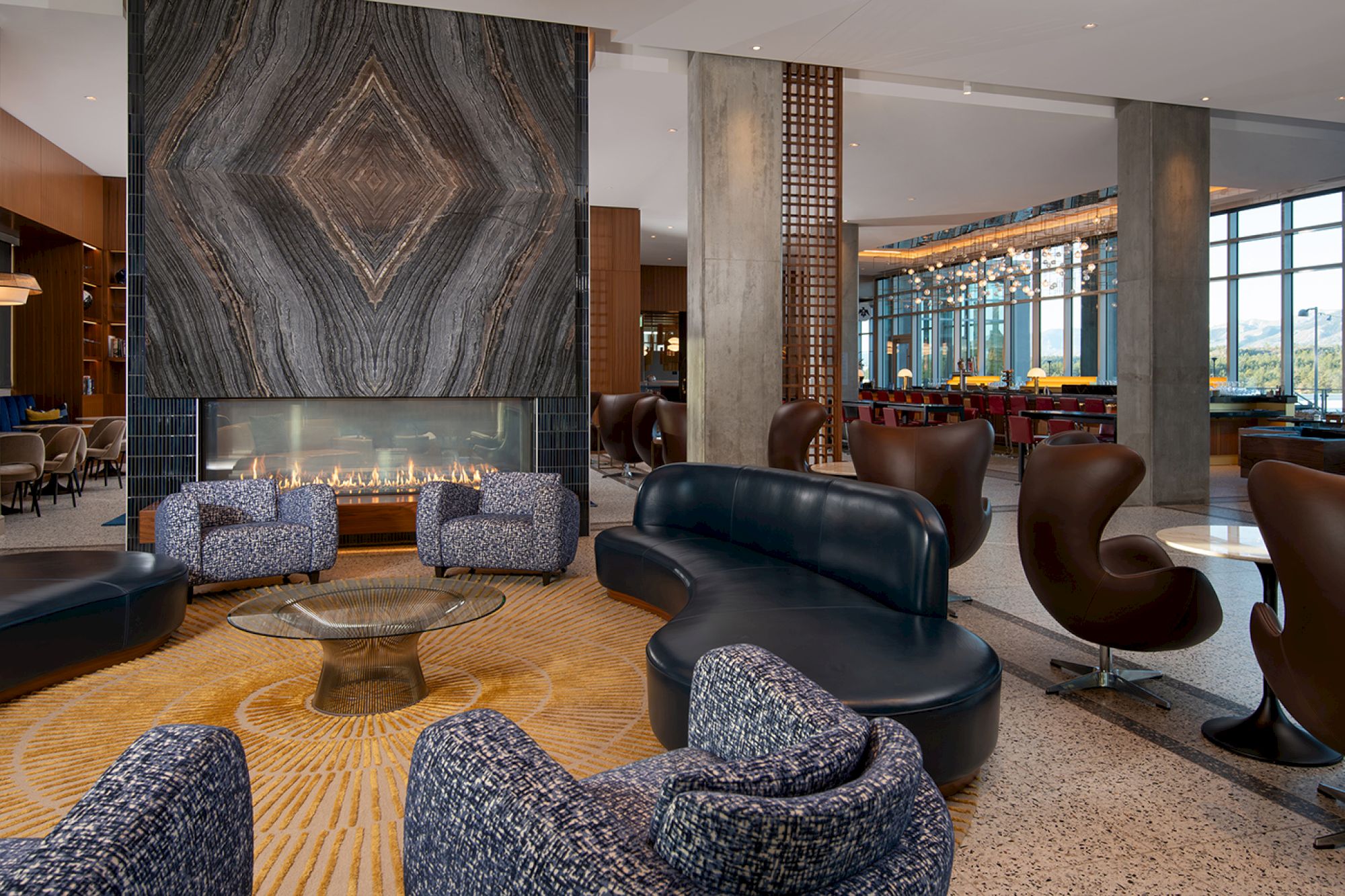 The image shows a modern lounge with plush seating, a geometric design fireplace, and large windows offering ample natural light.
