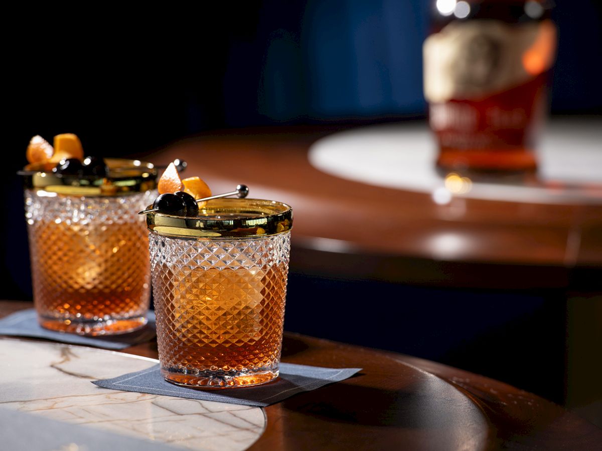 Two crystal glasses with an amber drink, garnished with orange peel and dark cherries, on a table with a bottle blurred in the background.