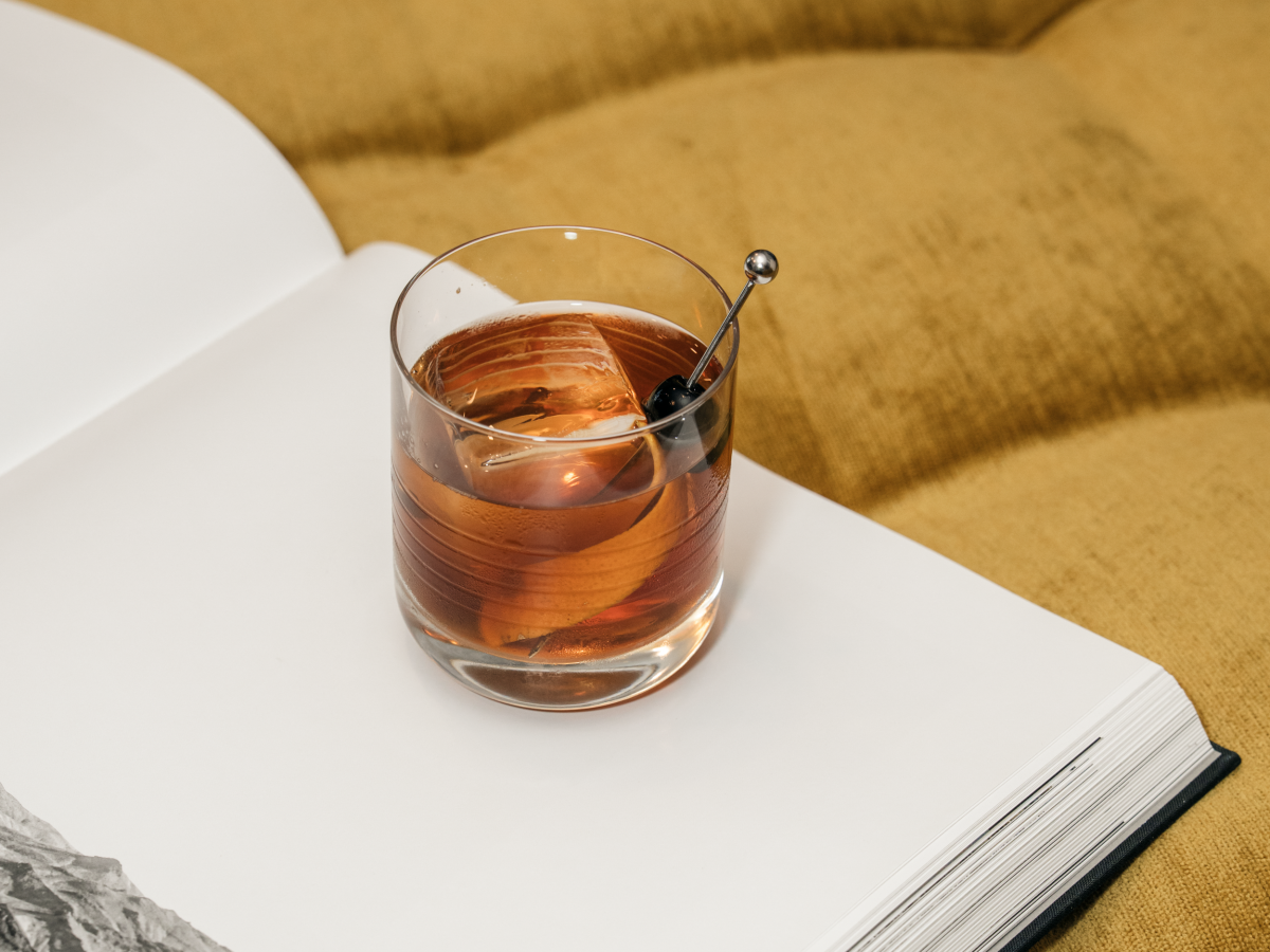 A glass of brown liquid with a stirrer is placed on an open book with black-and-white photos, resting on a yellow cushioned surface.