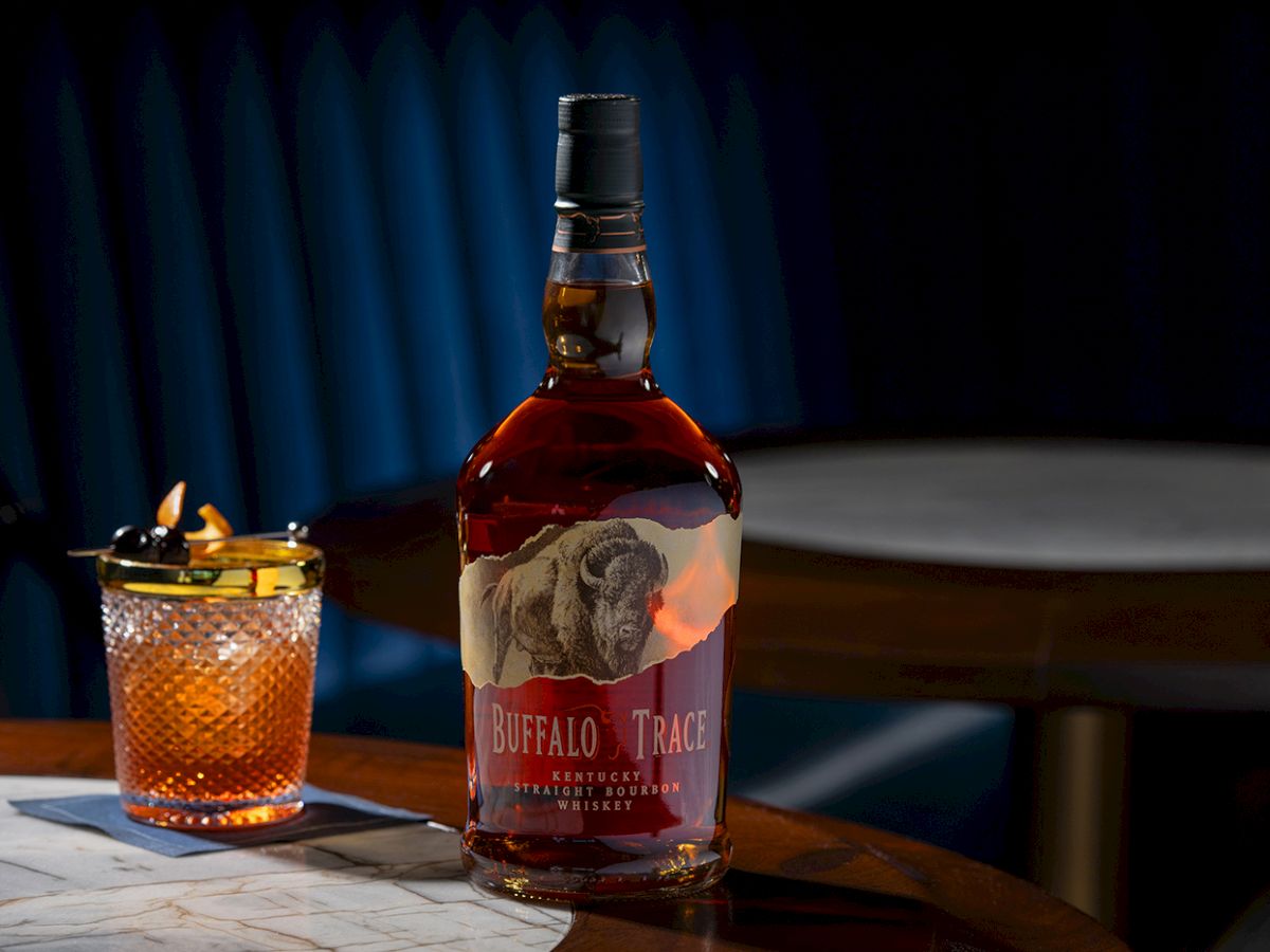 A bottle of Buffalo Trace bourbon and a glass with a garnish sit on a table in a dimly lit setting.