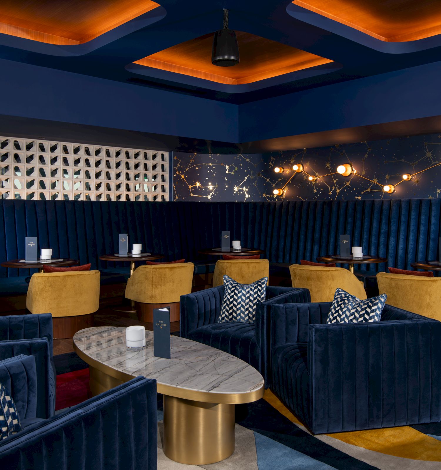 A stylish lounge area with blue velvet seating, yellow chairs, patterned cushions, marble tables, and decorative walls and lighting accents.